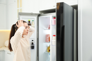 Emotional young woman near open refrigerator in kitchen