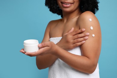 Photo of Young woman applying body cream onto shoulder on light blue background, closeup