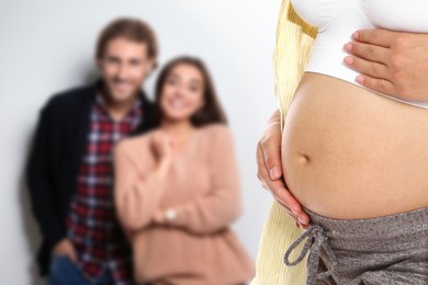 Surrogacy concept. Young pregnant woman and blurred view of happy couple on light background