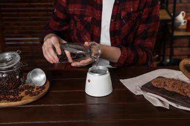 Brewing coffee. Woman pouring water into moka pot at wooden table indoors, closeup