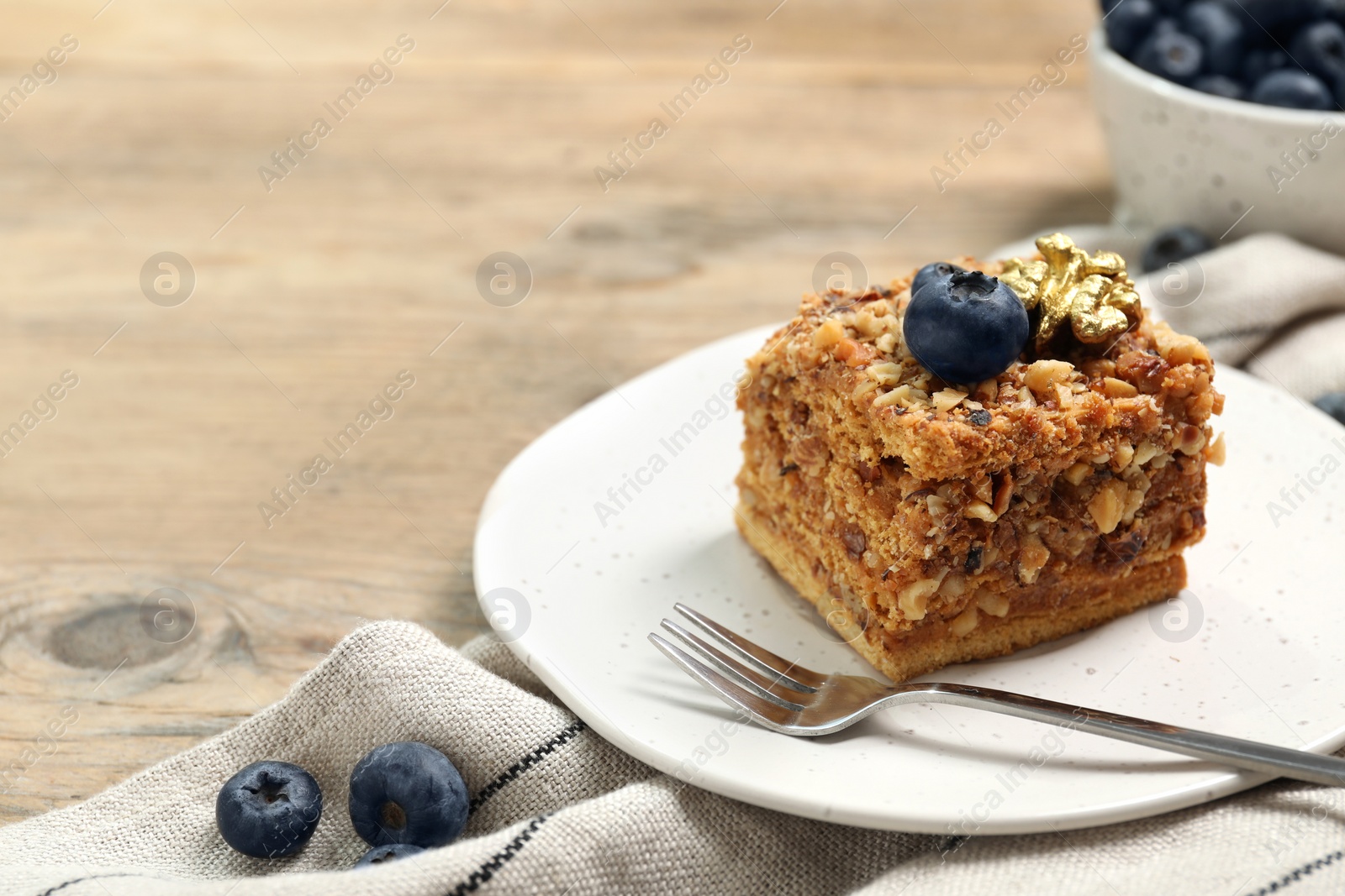 Photo of Piece of delicious layered honey cake with blueberries and nuts served on wooden table, closeup. Space for text