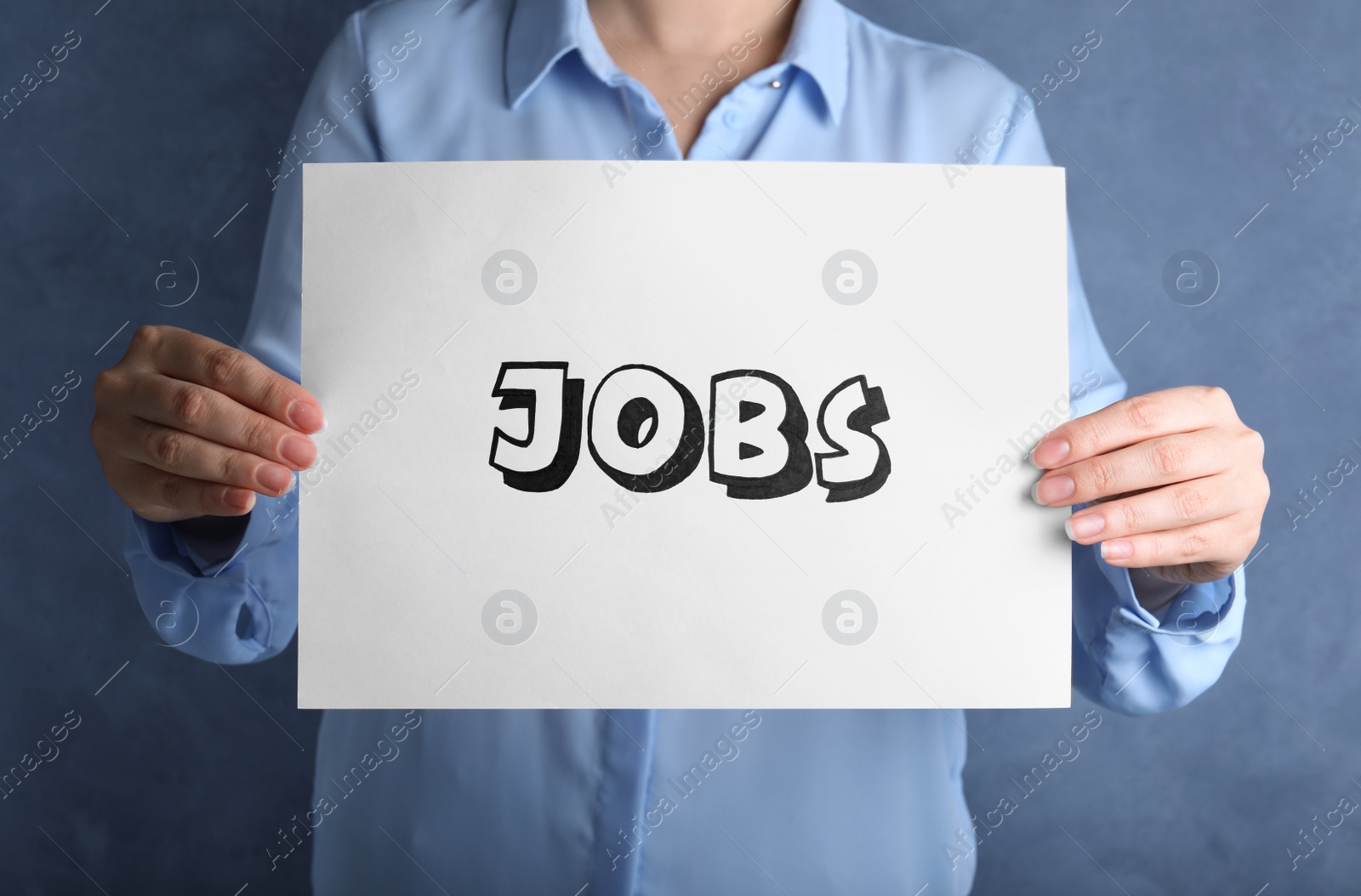 Photo of Woman holding sheet of paper with word JOBS on blue background, closeup. Career concept