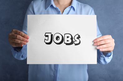 Photo of Woman holding sheet of paper with word JOBS on blue background, closeup. Career concept