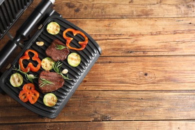 Electric grill with tasty meat steaks, rosemary and vegetables on wooden table, top view. Space for text