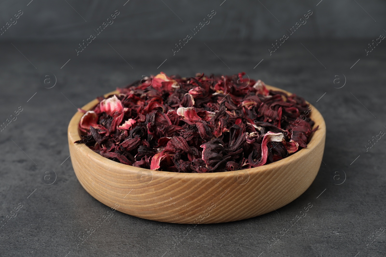 Photo of Hibiscus tea. Wooden bowl with dried roselle calyces on grey table, closeup