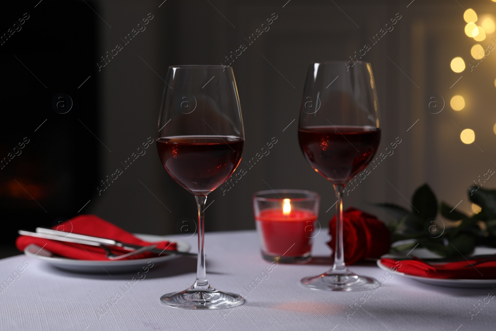 Photo of Romantic table setting with glasses of red wine, rose flower and burning candle against blurred lights, space for text