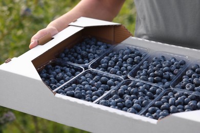 Photo of Man holding box with containers of fresh blueberries outdoors, closeup. Seasonal berries