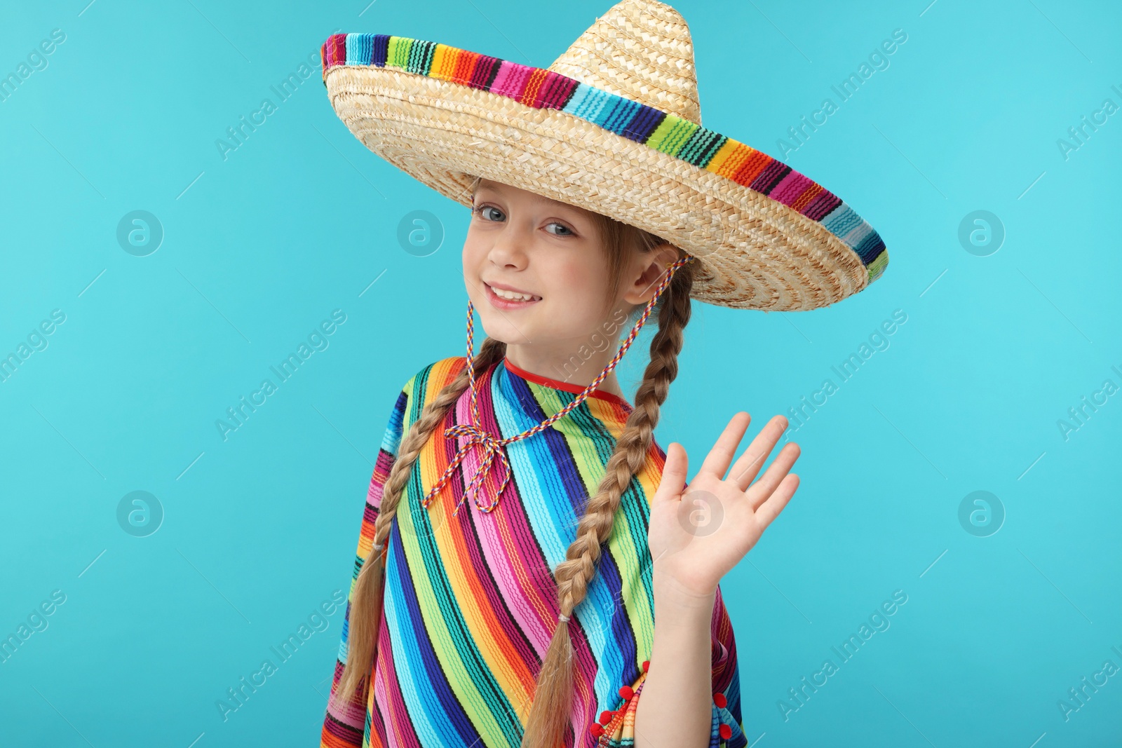 Photo of Cute girl in Mexican sombrero hat and poncho waving hello on light blue background