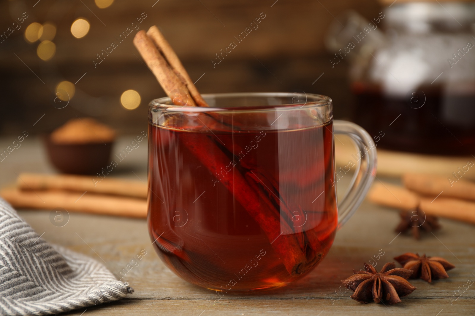 Photo of Glass cup of hot tea with aromatic cinnamon on wooden table against blurred lights