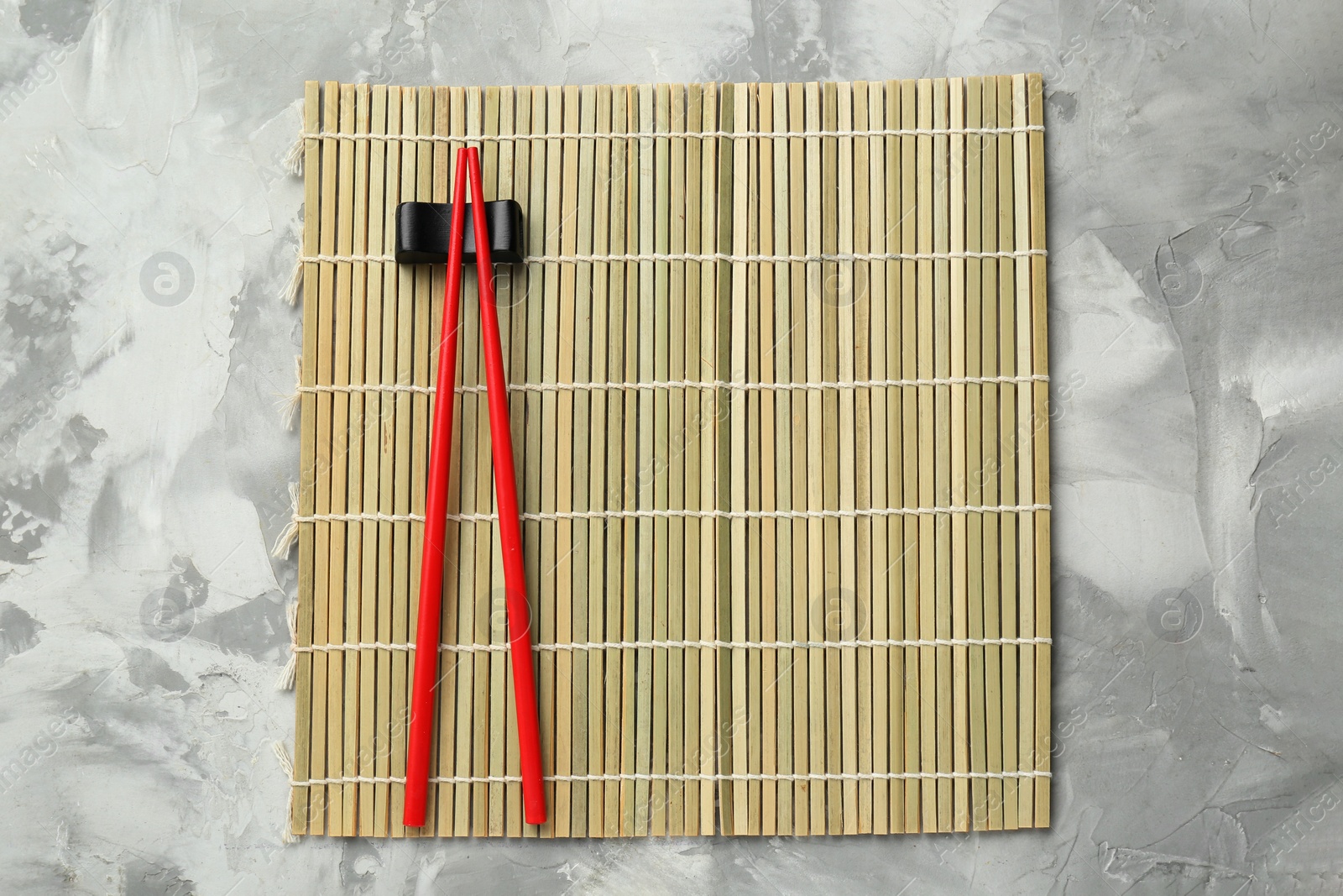 Photo of Bamboo mat with pair of red chopsticks and rest on grey textured table, top view