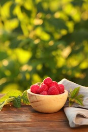 Photo of Tasty ripe raspberries in bowl and green leaves on wooden table outdoors, space for text