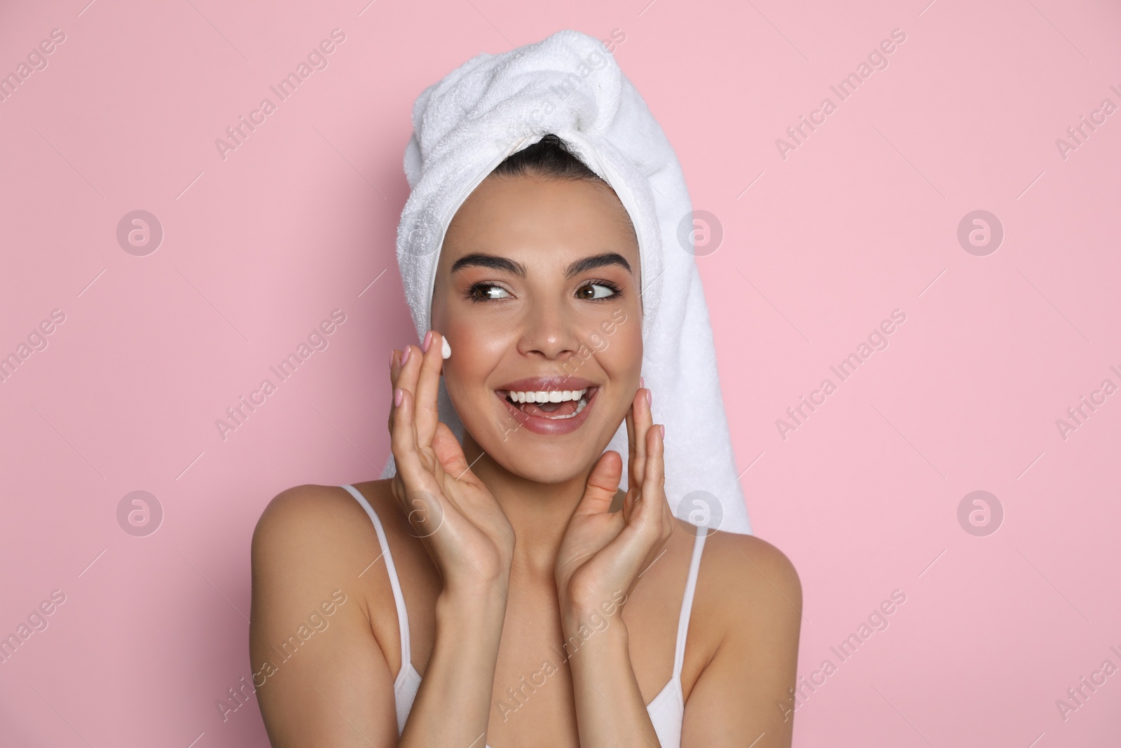 Photo of Beautiful young woman with towel applying cream on face against pink background