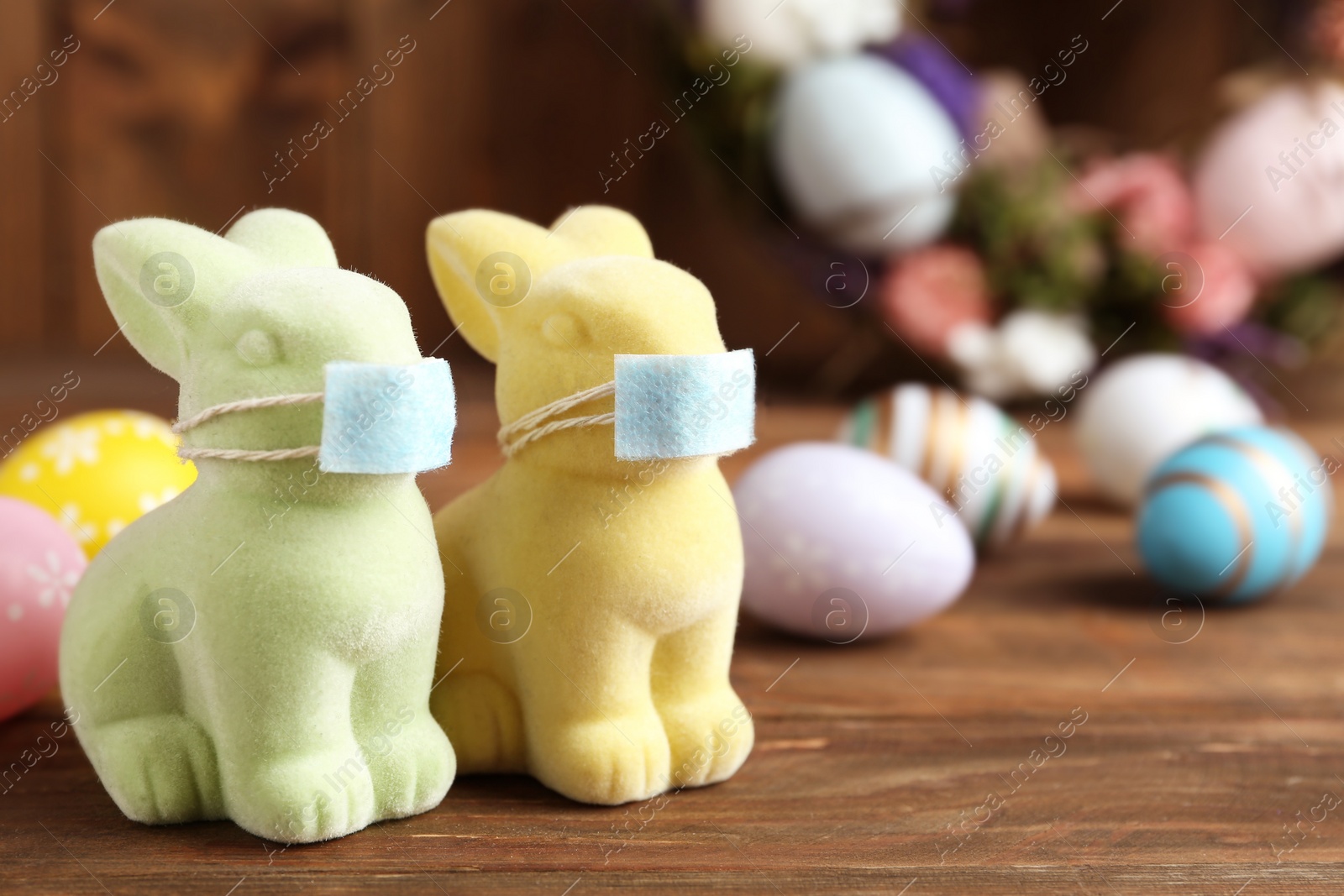 Photo of Cute bunny figures in protective masks near dyed eggs on wooden table, space for text. Easter holiday during COVID-19 quarantine