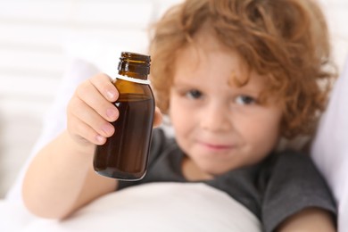 Photo of Cute boy holding cough syrup in bed, focus on bottle