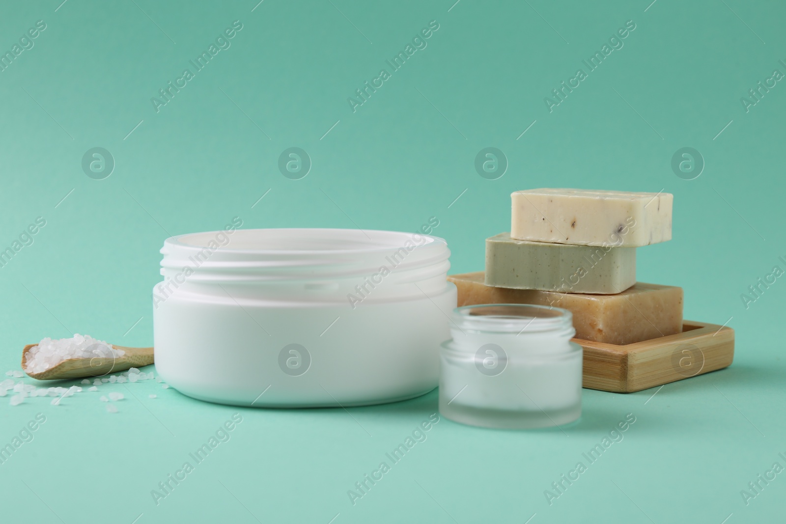 Photo of Moisturizing cream in open jars, sea salt and soap bars on turquoise background. Body care products