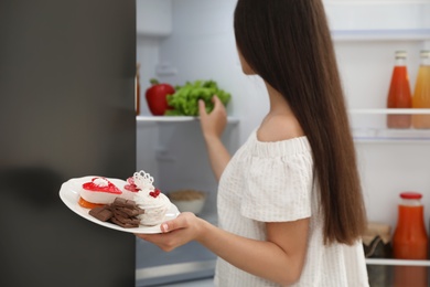 Photo of Concept of choice between healthy and junk food. Woman holding plate with sweets near refrigerator in kitchen, focus on hand