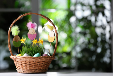 Photo of Wicker basket with colorful Easter eggs, wooden figures and flowers on white wooden table against blurred green background. Space for text