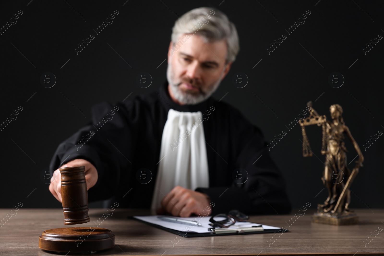 Photo of Judge with gavel and papers sitting at wooden table against black background, selective focus