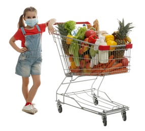 Photo of Little girl in medical mask with shopping cart full of groceries on white background