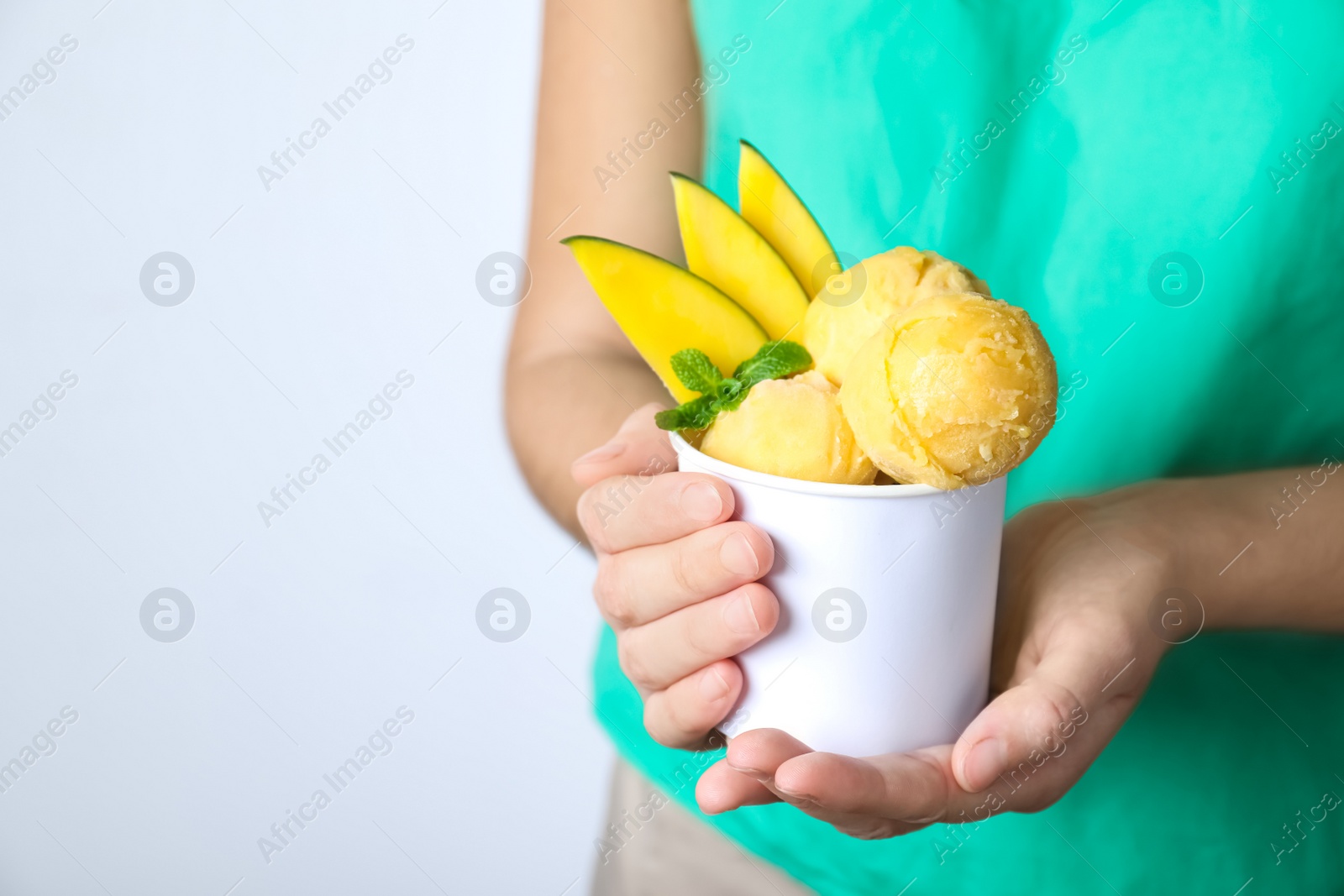 Photo of Woman holding bowl full of delicious ice cream and mango slices on light grey background, closeup. Space for text