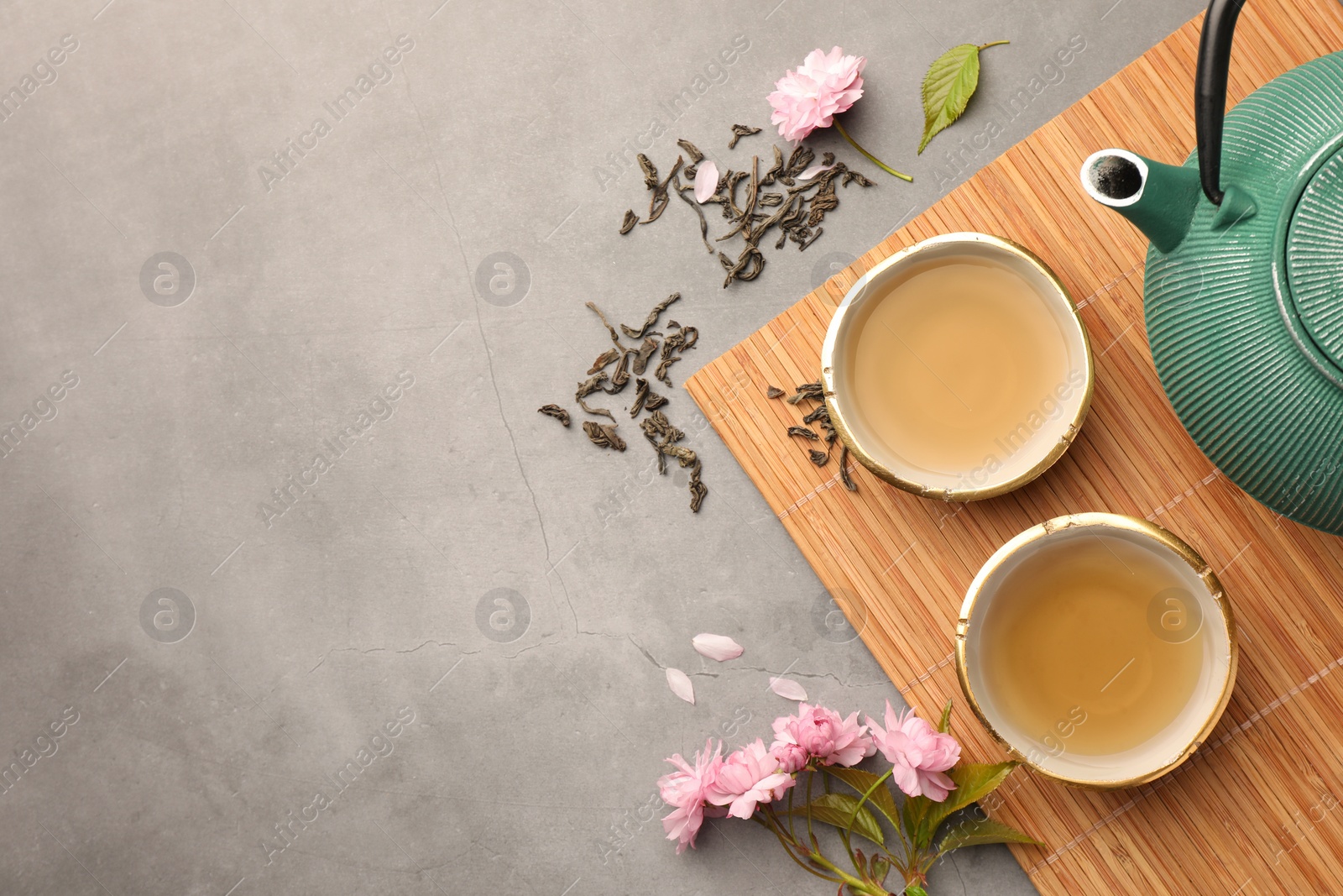 Photo of Traditional ceremony. Cups of brewed tea, teapot and sakura flowers on grey table, flat lay with space for text