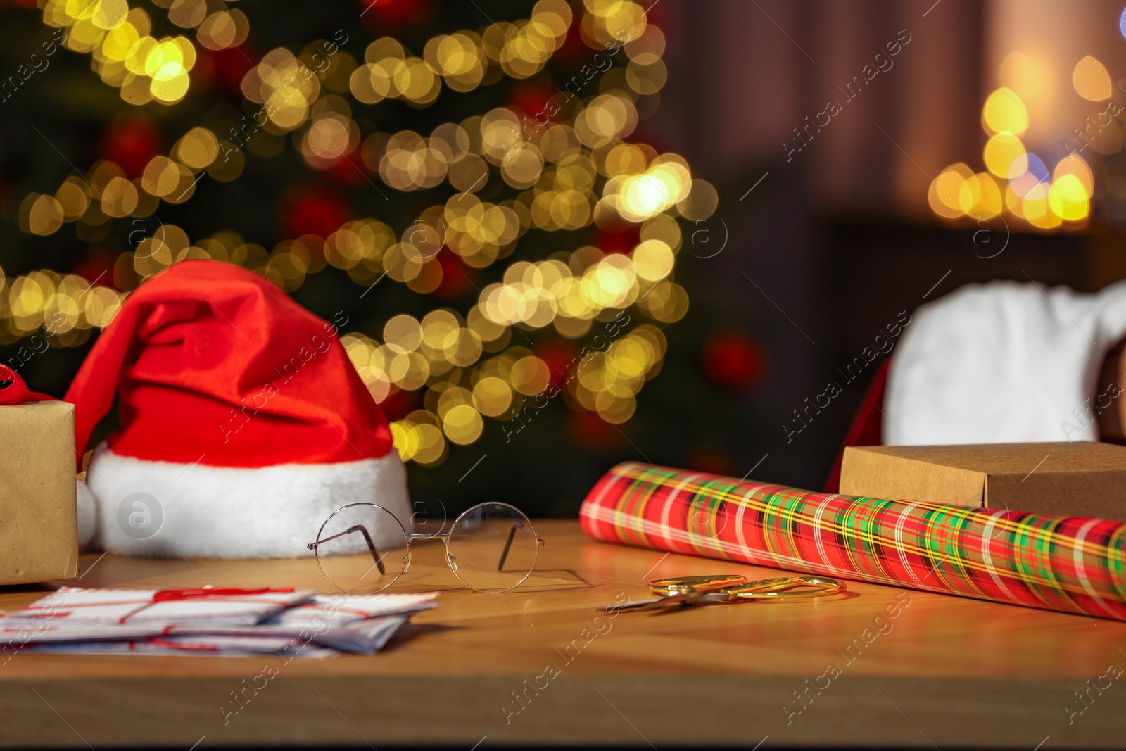 Photo of Santa's Claus workplace. Gift box, wrapping paper, letters and hat on table in room with Christmas tree