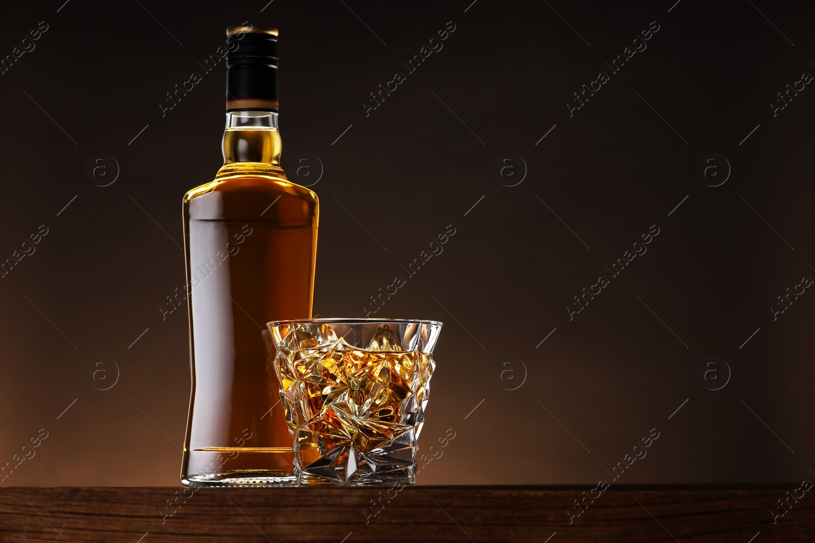 Photo of Whiskey with ice cubes in glass and bottle on wooden table against brown background, space for text