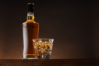 Photo of Whiskey with ice cubes in glass and bottle on wooden table against brown background, space for text