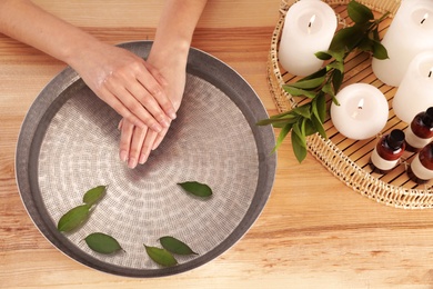 Photo of Woman soaking her hands in bowl with water and leaves on wooden table, top view. Spa treatment