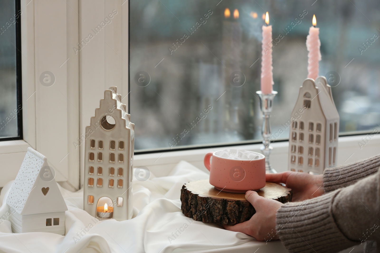 Photo of Woman holding cup of delicious drink near windowsill with house shaped lanterns indoors, closeup