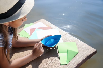 Little girl making paper boats on wooden pier near river