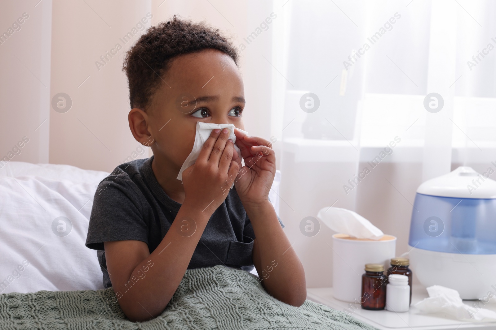 Photo of African-American boy with tissue blowing nose in bed indoors, space for text. Cold symptoms