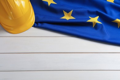 Photo of European Union flag and yellow hard hat on white wooden table, flat lay. Space for text