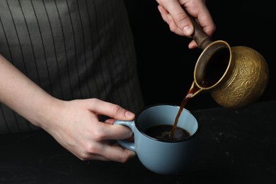 Photo of Turkish coffee. Woman pouring brewed beverage from cezve into cup at black table, closeup