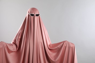 Photo of Glamorous ghost. Woman in pink sheet with sunglasses on light grey background, space for text