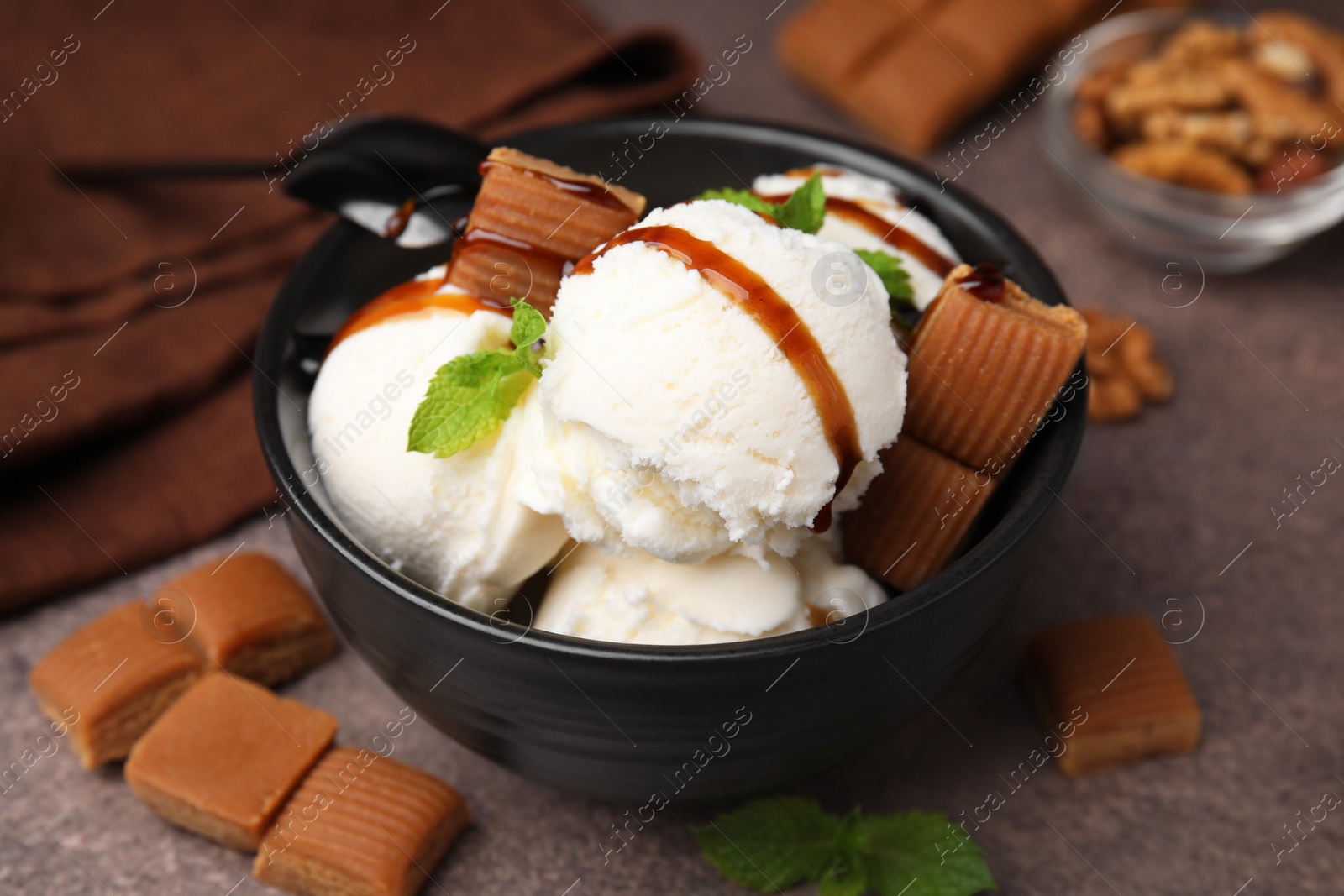 Photo of Bowl of tasty ice cream with caramel sauce, candies and mint on brown table, closeup