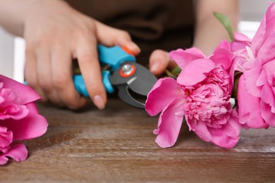 Photo of Woman trimming beautiful pink peonies with secateurs at wooden table, selective focus
