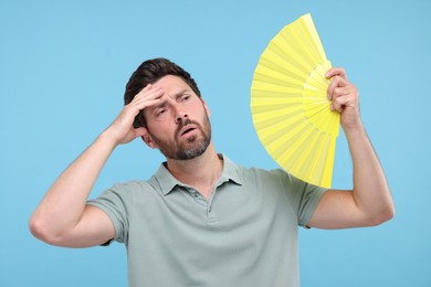 Unhappy man with hand fan suffering from heat on light blue background