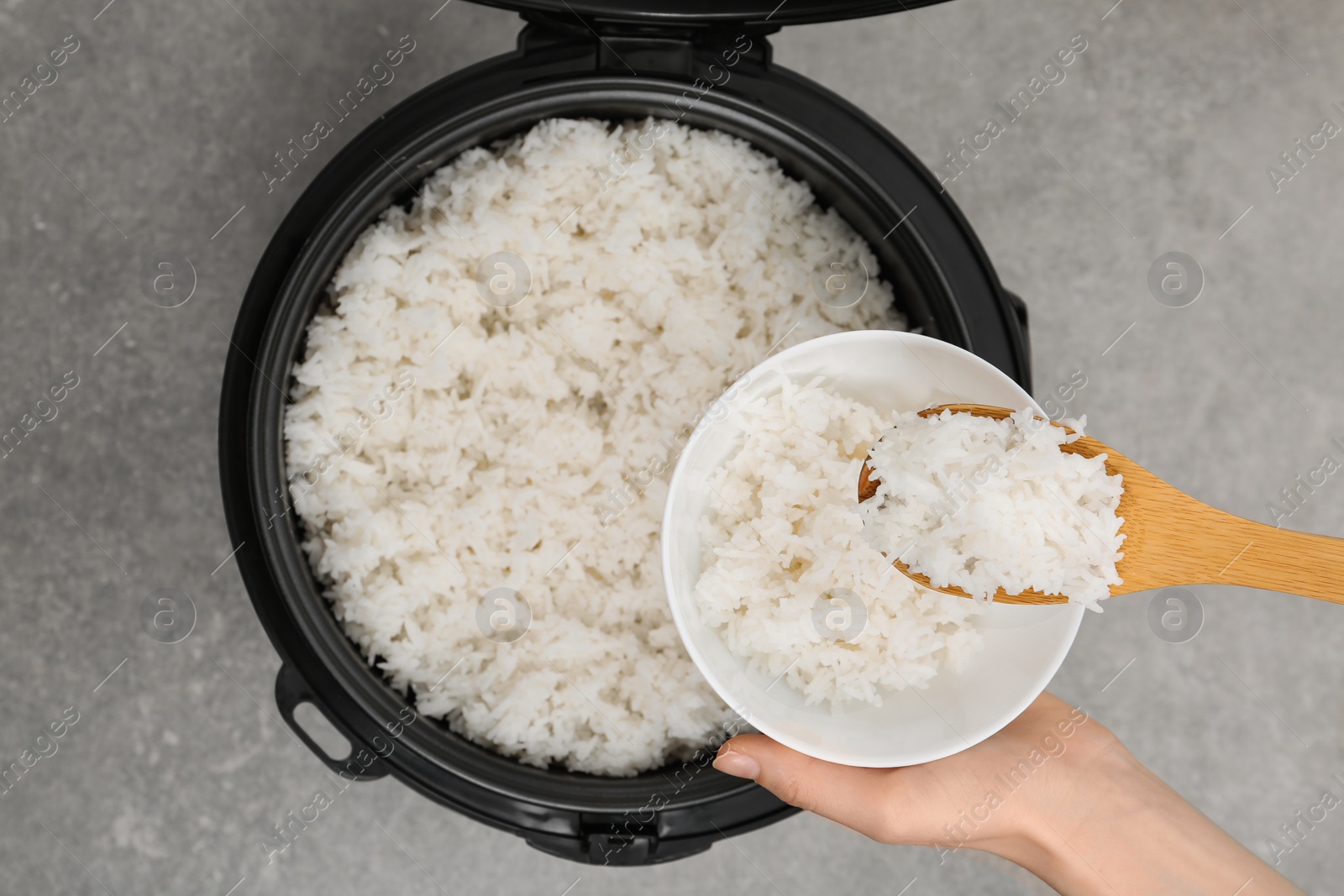 Photo of Woman putting boiled rice into bowl from cooker on grey background, top view