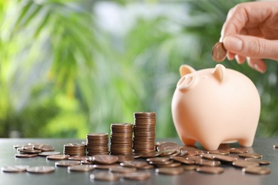 Photo of Woman putting coin into piggy bank at grey table against blurred green background, closeup. Space for text