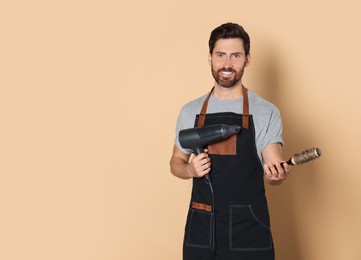 Photo of Smiling hairdresser in apron holding dryer and brush on light brown background, space for text
