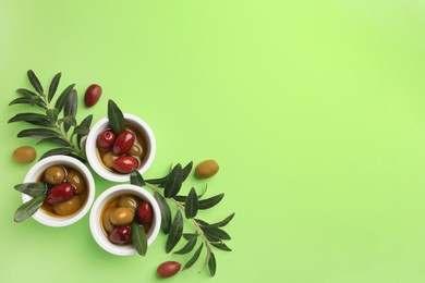 Bowls with different ripe olives and leaves on light green background, flat lay. Space for text