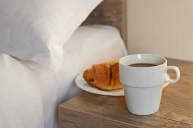 Photo of Cup of morning coffee and croissant on wooden night stand near bed indoors