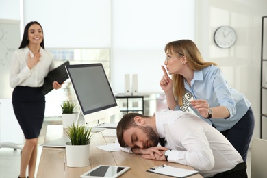 Photo of Young woman sticking paper fish to colleague's back while he sleeping in office. Funny joke