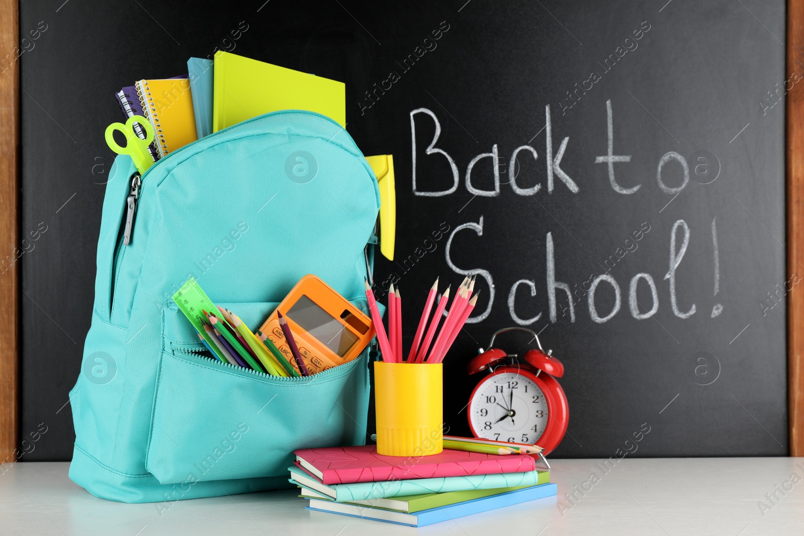 Photo of Bright backpack with school stationery on white table near black chalkboard. Back to School