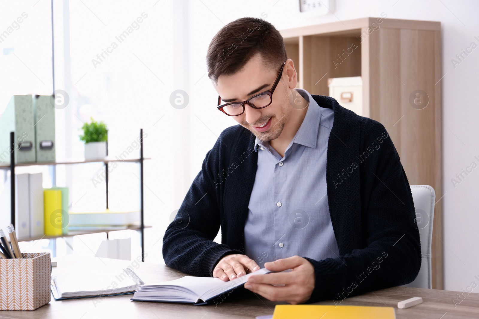 Photo of Young male teacher with book sitting at table in classroom