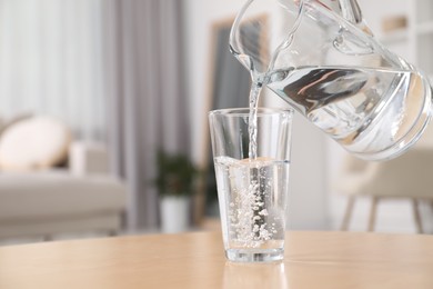 Pouring fresh water from jug into glass at table against blurred background, closeup. Space for text