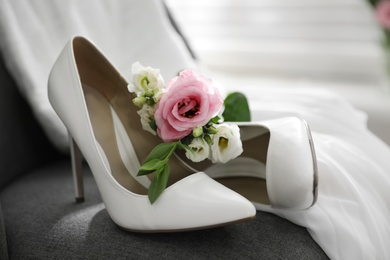 Photo of Pair of white high heel shoes, flowers and wedding dress on chair indoors, closeup