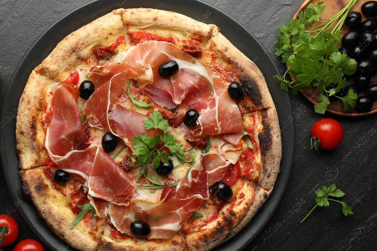 Photo of Tasty pizza with cured ham, olives, tomatoes and parsley on black table, top view