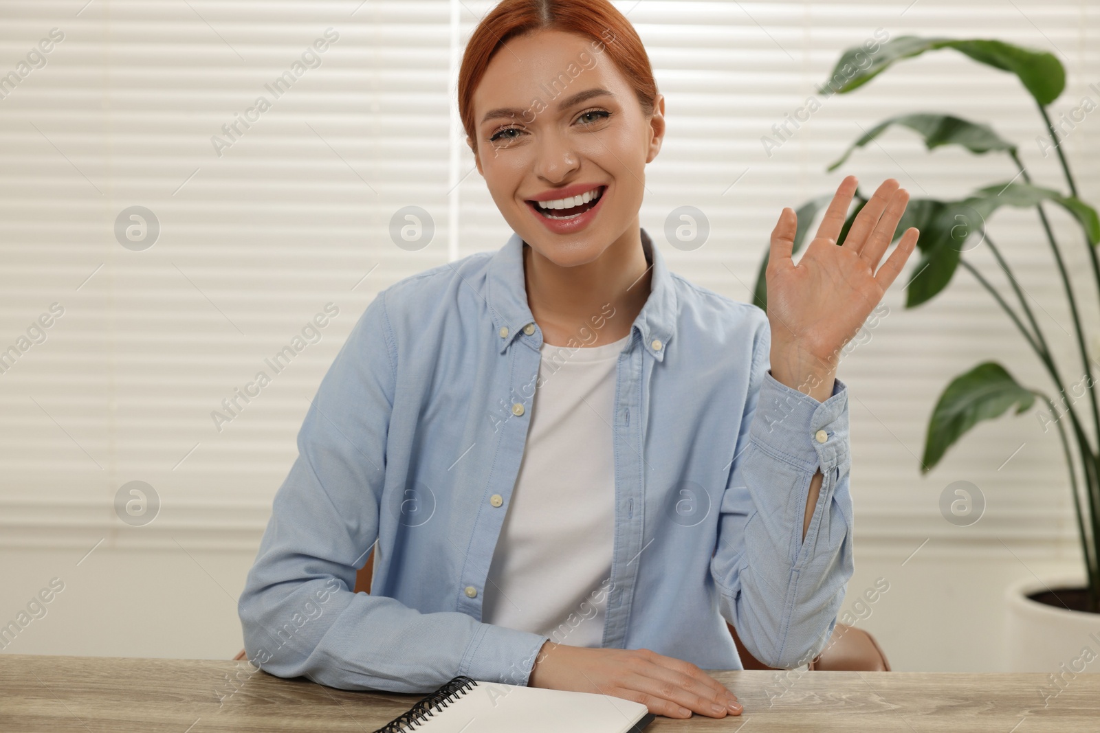 Photo of Young woman waving hello during video chat at wooden table indoors, view from web camera